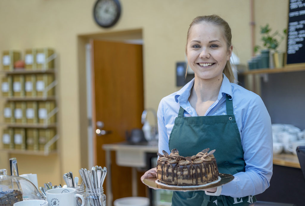 Vaaleansiniseen kauluspaitaan ja vihertävään esiliinaan pukeutunut poninhäntäpäinen nainen pitää käsissään lautasta, jossa on upea suklainen täytekakku. Naisen vieressä näkyy kahvikuppeja ja astia, jossa on lusikoita pystyssä. Takana näkyy hylly, jossa on laaja teevalikoima sekä ovi, jonka yläpuolella on kello. Naisen takana on myös toinen hylly, jossa on viherkasvi sekä lisää kahvikuppeja.