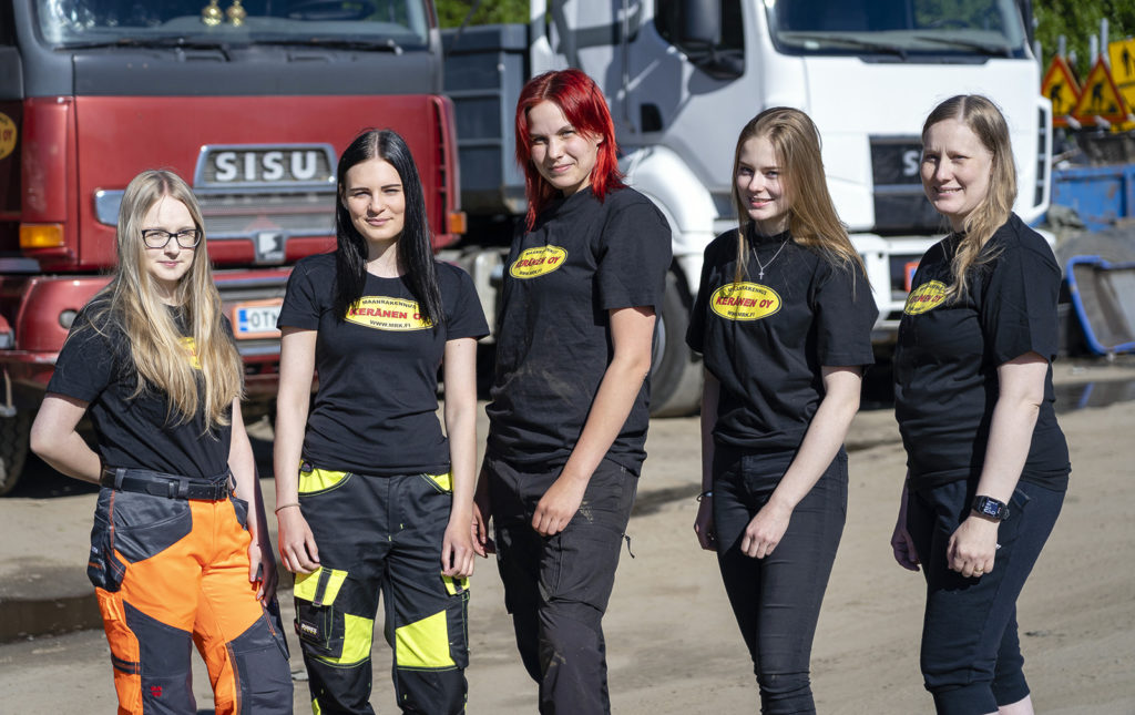 Viisi nuorta naista seisoo rivissä kuorma-autojen edessä. Jokaisella naisella on musta t-paita päällään, jossa on keltainen soikea alue, jossa lukeen Maanrakennus Keränen Oy.
