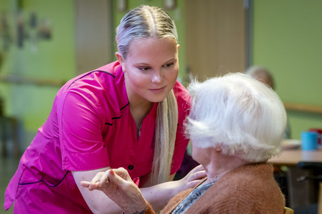 Pinkkiin asuun pukeutunut hoitaja jututtaa vanhusta.