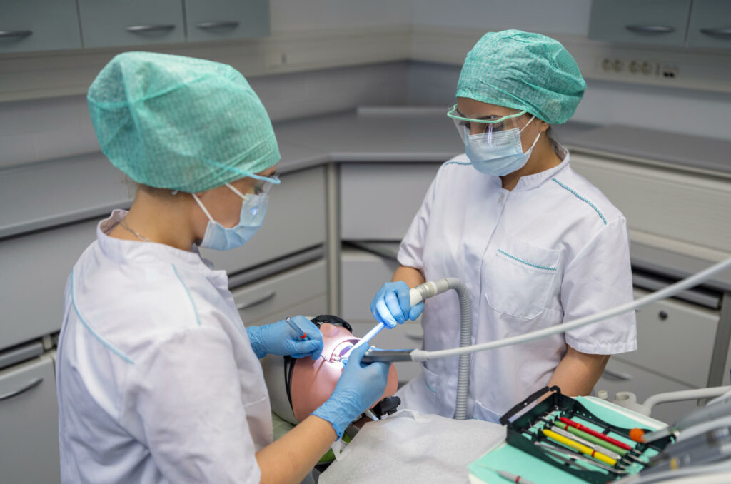Kaksi suojavaatteisiin pukeutunutta henkilöä työskentelee hammashoitolan hoitotuolin ääressä.