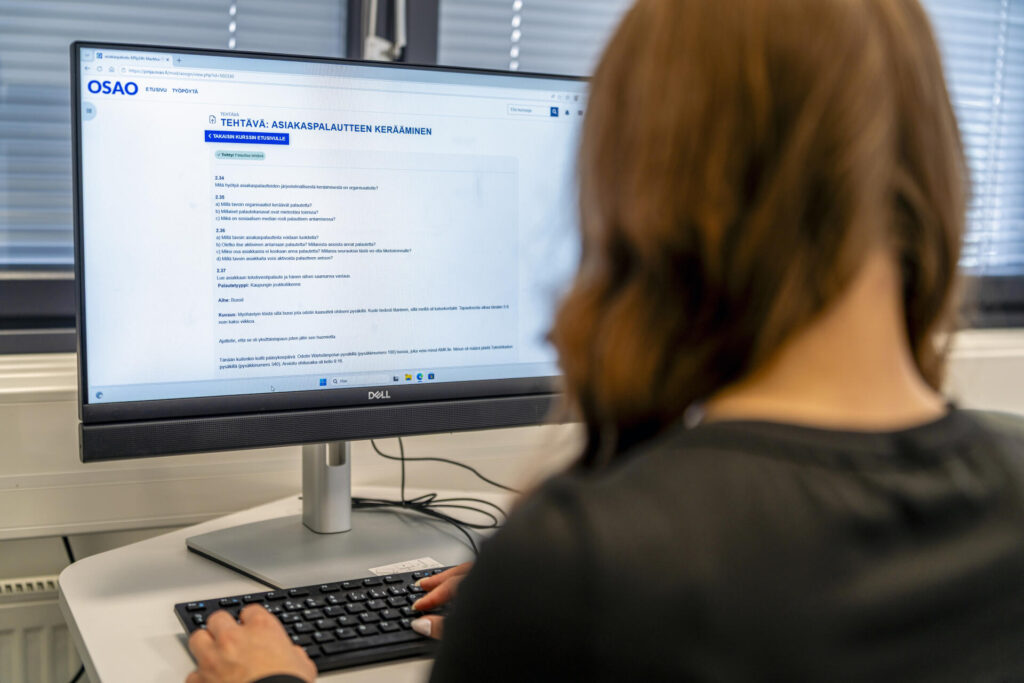 Henkilö istuu tietokoneen ääressä työpöydällä. Näytöllä on tekstiä ja otsikko "Tehtävä: Asiakaspalautteen kerääminen". Näyttöön on liitetty näppäimistö ja hiiri.