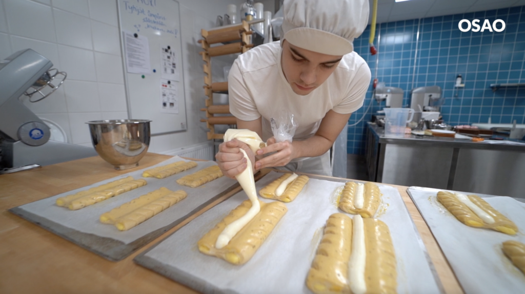 Leipuri-kondiittoriopiskelija pursottaa kreemiä pullapitkojen keskustaan.