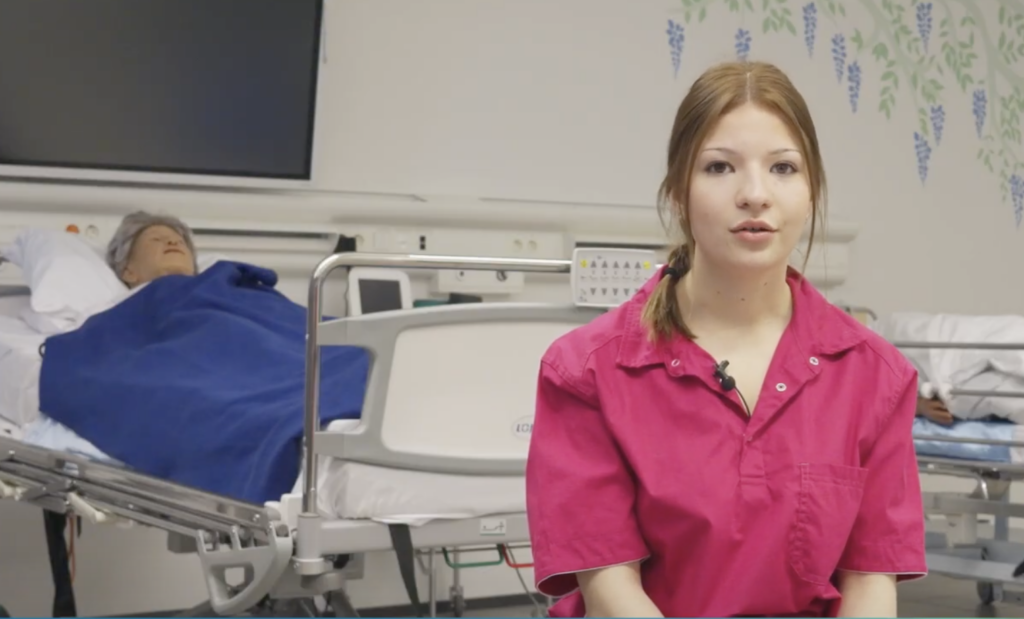 Nainen istuu sairaalassa potilashuoneessa. Taustalla on sairaalasänky, jossa on siniseen peittoon peitetty nukke. Naisella on yllään vaaleanpunainen paita ja hän katsoo kameraan.