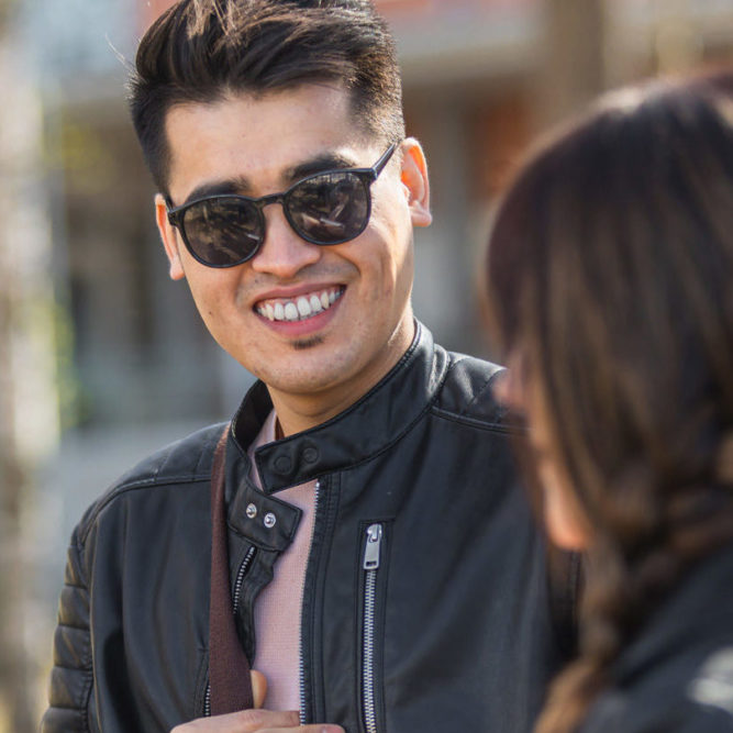 Dark-haired man with sunglasses smiling to a wdark-haired woman in front of him. There's trees and a building on the background.