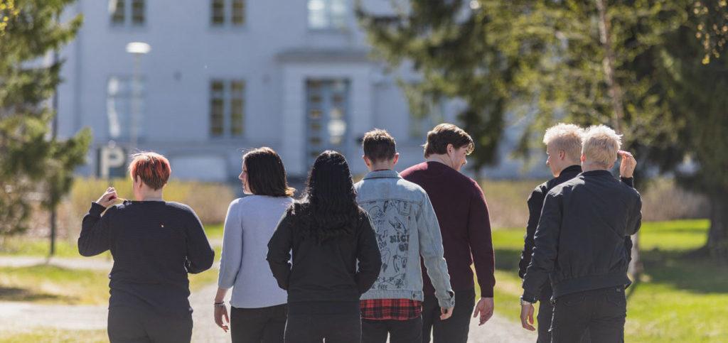 A group of students are walking away from the camera towards a white building. There's greenery by the side.