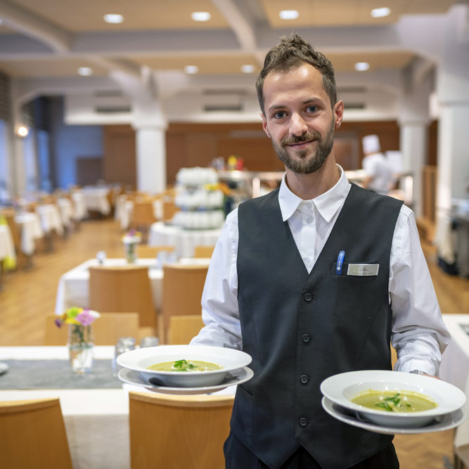 Mies valkoisessa paidassa ja tummassa liivissä pitelee käsisään lautasia, joilla on keittoannokset. Taustalla ravintolasali.