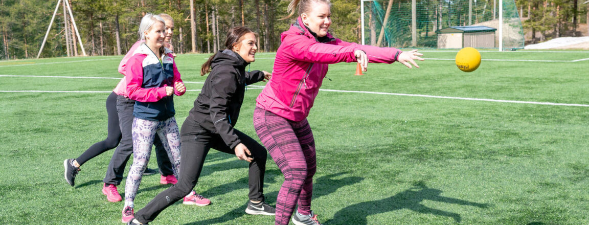 Ryhmä naisia seisoo jonossa vihreällä jalkapallokentällä. Jonon ensimmäisen nainen heittää juuri keltaisen pallon eteenpäin. Jonossa olevat muut naiset kurkkivat jonossa edellä olevien selkien takaa.