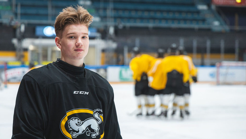 Nuorimies seisoo jääkiekkokaukalon jäällä. Hänellä on päällään musta paita, jossa on rinnassa keltainen soikio, jonka keskellä on valkoinen kärpän kuva. Miehen takana seisoo ryhmä keltapaitaisia jääkiekkoilijoita.