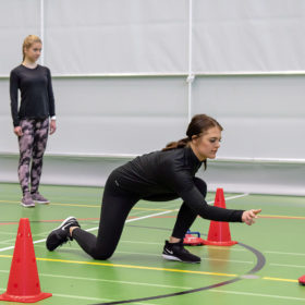 Tummaan asuun pukeutunut tummahiuksinen nainen lähettänyt punaisen curling-kiekon matkaan punaisten tötsien välistä. Naisen toinen polvi on maassa ja toinen jalka koukussa edessä. Taustalla on kolme muuta naista seuraamassa suoritusta.
