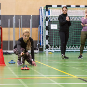 Vaaleahiuksinen nainen on kyykyssä punaisten tötsien välissä. Hänen toinen kätensä on ojennettuna eteen ja hän on lähettänyt punaisen curling-kiven liukumaan pitkin liikuntasalin lattiaa. Takana on muita henkilöitä seuraamassa suoritusta. Takana näkyy myös futsal-maalit.