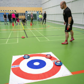 Lyhythihaiseen paitaan ja shortseihin pukeutunut mies seuraa kuinka punainen curling-kivi liukuu kohti curling-kentän maalipesää. Pesässä on jo kaksi sinistä kiveä ja yksi punainen kivi. Kauempana seinän vieressä seisoo isompi joukko ihmisiä, jotka ovat pelaamassa liikuntasalissa curlingia.