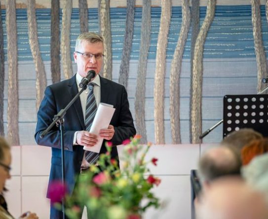 Kuntayhtymäjohtaja-rehtori Jarmo Paloniemi pitää puhetta juhlassa. Hän puhuu mikrofoniin ja pitelee paperikääröä kädessään.