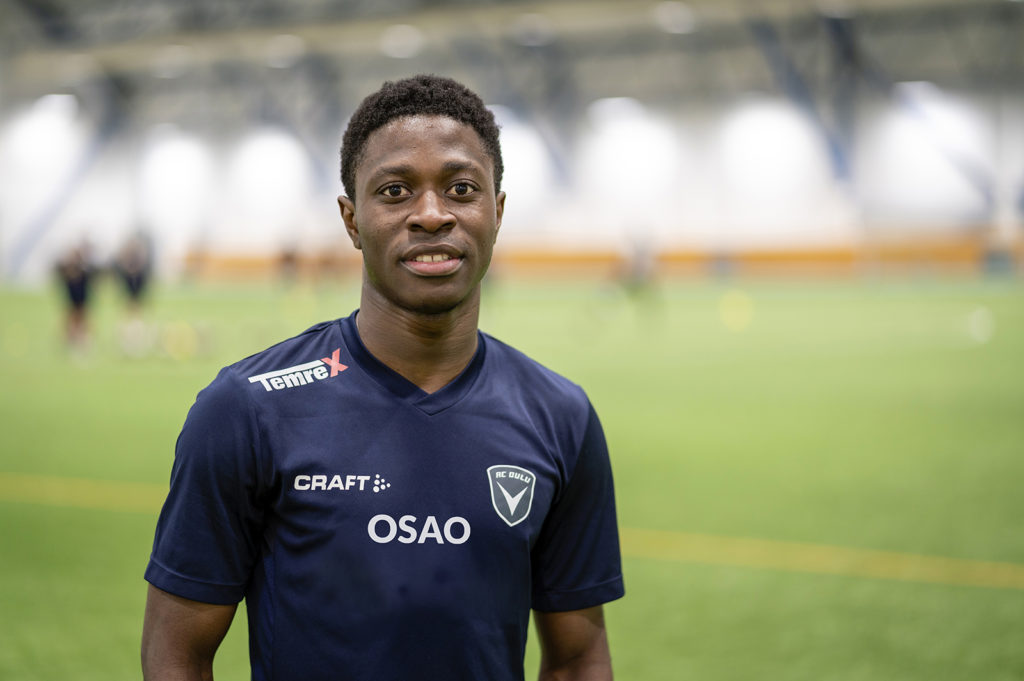 Tummaihoinen mies tummansinisessä urheilupaidassa seisoo jalkapallohallin tekonurmen edessä. Miehen paidassa on edessä OSAOn logo.