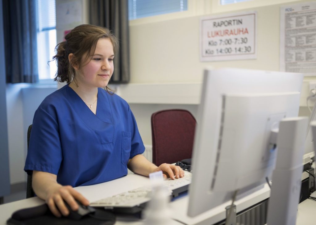 Siniseen lyhythihaiseen terveydenhuollon henkilöstön työpaitaan pukeutunut nuori nainen istuu tietokoneen ääressä. Hänen kätensä ovat näppäimistöllä ja katse tiiviisti näytössä.