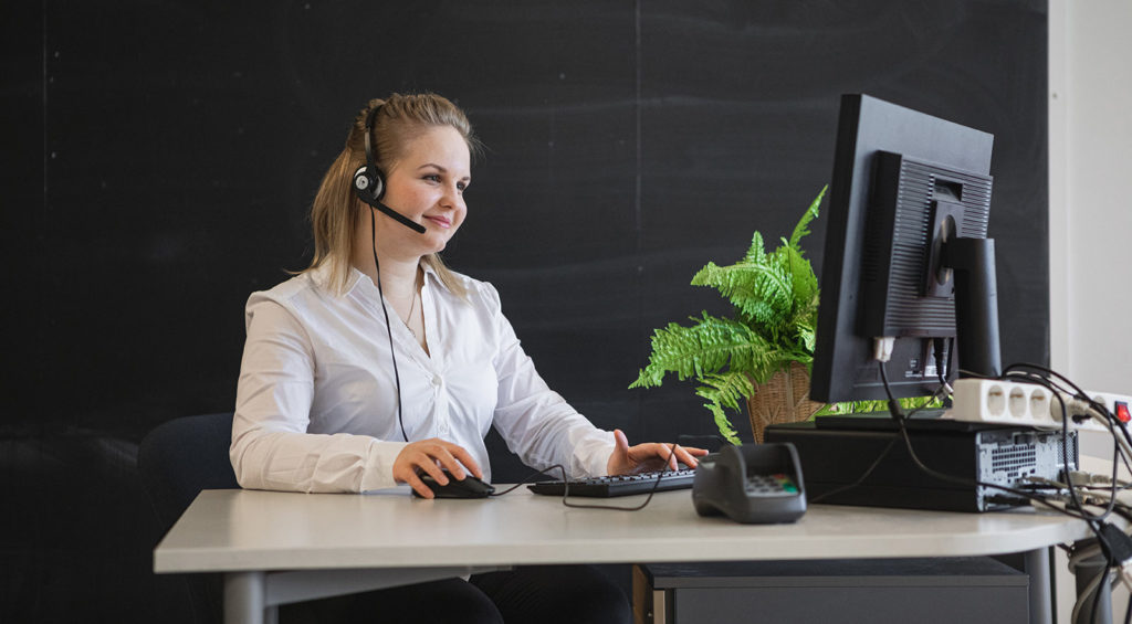 Vaaleahiuksinen nuori nainen istuu tietokoneen ääressä. Naisella on kuuloke-mikrofoni headset päässään. Pöydällä on tietokoneen rinnalla kassapääte ja viherkasvi.