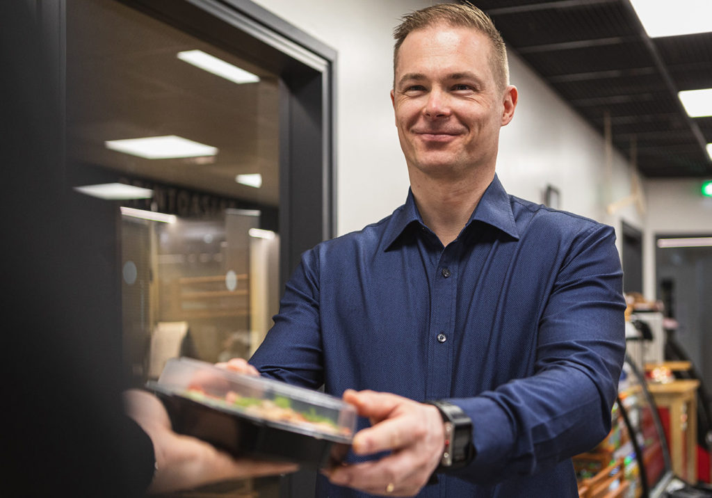 Hymyilevä mies sinisessä kauluspaidassa ojentaa tai ottaa vastaan rasiaa, jonka sisällä on salaattia.