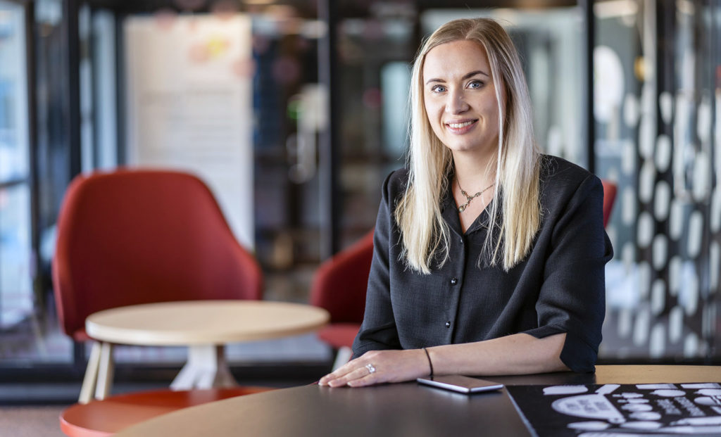 Vaalea, pitkähiuksinen nainen istuu pöydän ääressä. Naisella on tummanharmaa paita päällä. Takana näkyy punainen nojatuoli ja pieni pyöreä pöytä.