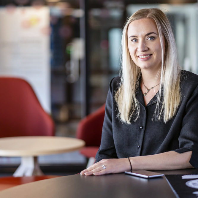 Vaalea, pitkähiuksinen nainen istuu pöydän ääressä. Naisella on tummanharmaa paita päällä. Takana näkyy punainen nojatuoli ja pieni pyöreä pöytä.