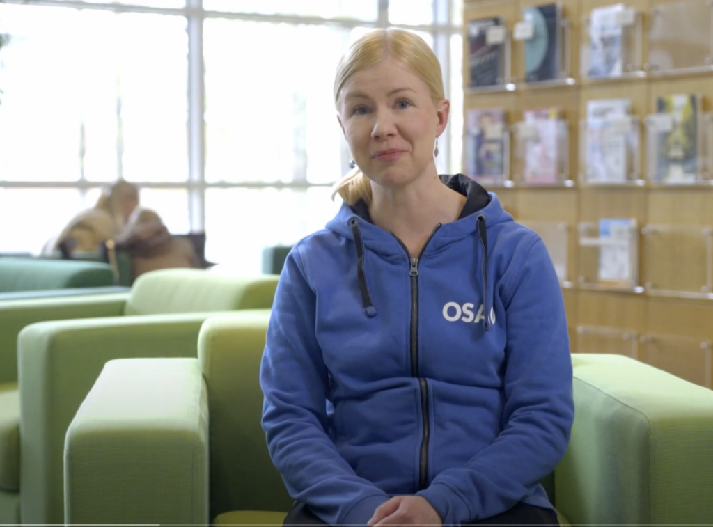 Nainen istuu vihreällä sohvalla sinisessä hupparissa, jossa on teksti 'OSAO'. Hänen takanaan on kirjahylly ja muita ihmisiä istumassa.