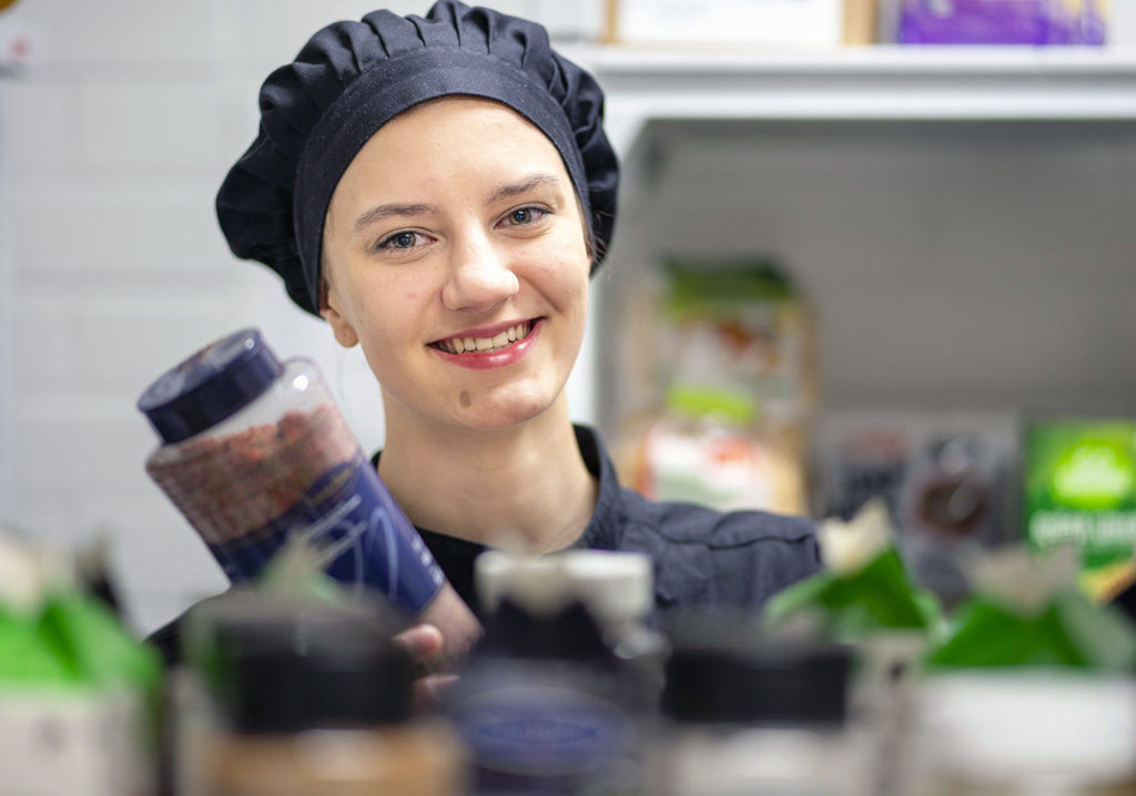 Mustaan kokinhattuun pukeutunut nainen on maustehyllyn takana ja pitää kädessään sinikantista isoa maustepurkkia, jossa on vaaleanruskeaa jauhetta.