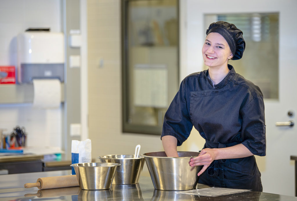 Mustaan kokinhattuun ja kokin työtakkiin pukeutunut nainen seisoo teräksisen työpöydän vieressä. Pöydällä on kolme teräksistä kulhoa. Naisen käsi on työnnetty suurimpaan kulhoista. Toisella kädellä hän pitää kulhon reunasta kiinni.
