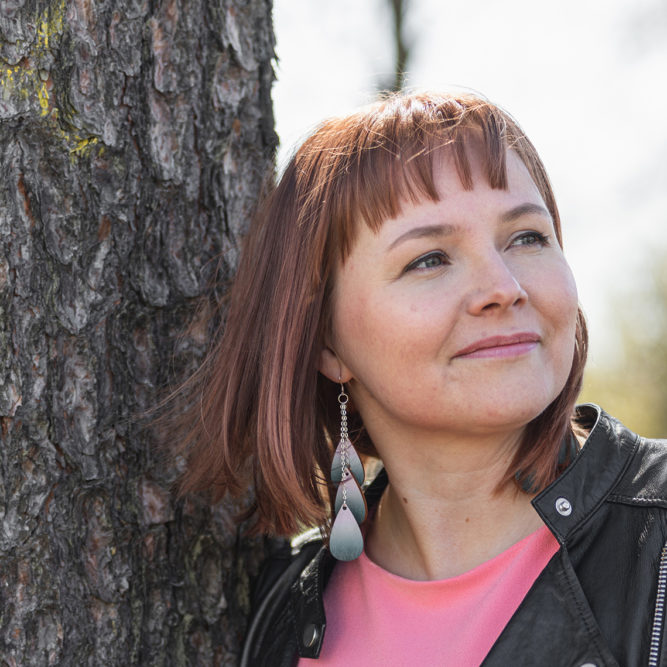 Punertavahiuksinen nainen nojaa paksuun puunrunkoon ja katsoo tulevaisuuteen.
