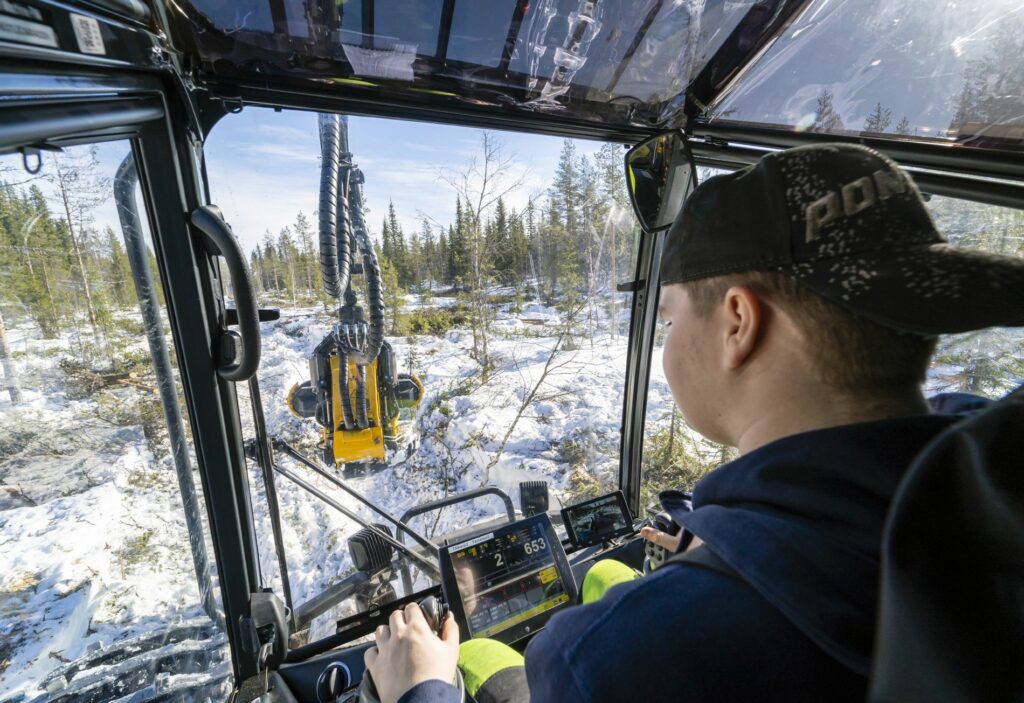 Lippalakkipäinen mies istuu metsäkoneen hytissä ja nostaa puita metsäkoneen kauhalla.