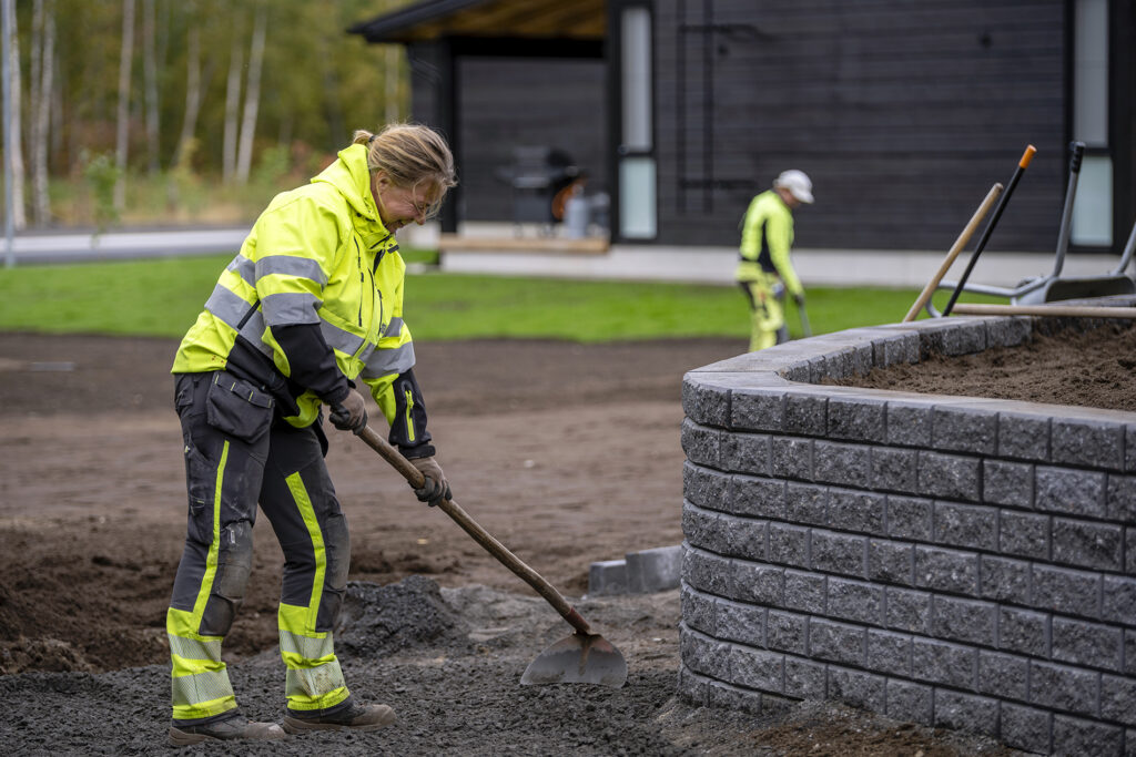 Nainen keltaisissa työvaatteissa levittää multaa juuri rakennetulle virheralueelle. Edessä kivimuuri.