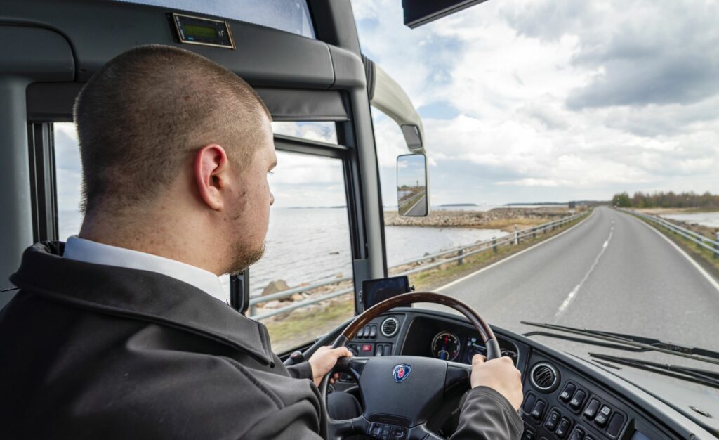 Mies ajaa linja-autoa. Edessä näkyy tie ja meri.