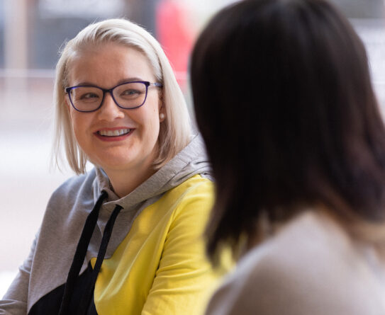Vaaleahiuksinen nainen istuu ja katsoo hymyillen kohti edessä, selin kameraan istuvaa tummahiuksista henkilöä.