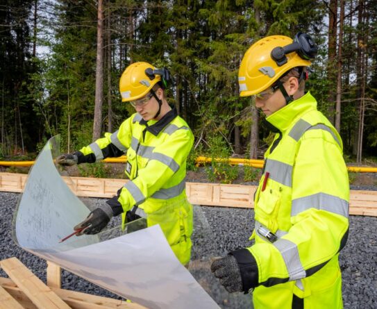 Kaksi talonrakentajaa tarkastelevat rakennuspiirustuksia. henkilöillä on työasut päällä ja takana kesäinen mäntymetsä.