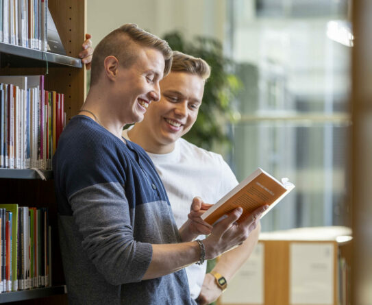 Kaksi nuorta miestä kirjastossa katsomassa toisen miehen käsissä oleva kirjaa.