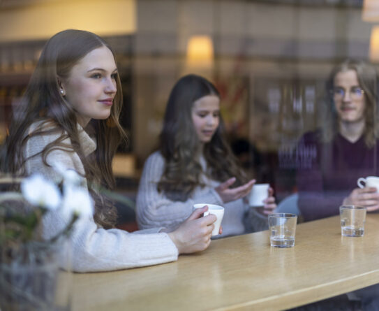 Kolme nuorta istuu kahvilassa ikkunan ääressä olevassa pöydässä, käsissään kahvikupit.