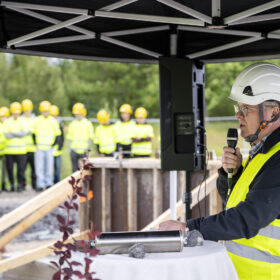 Mies puhuu mikrofoniin työmaalla ja yleisö kuuntelee taustalla. Kaikilla on turvavarusteet päällä.