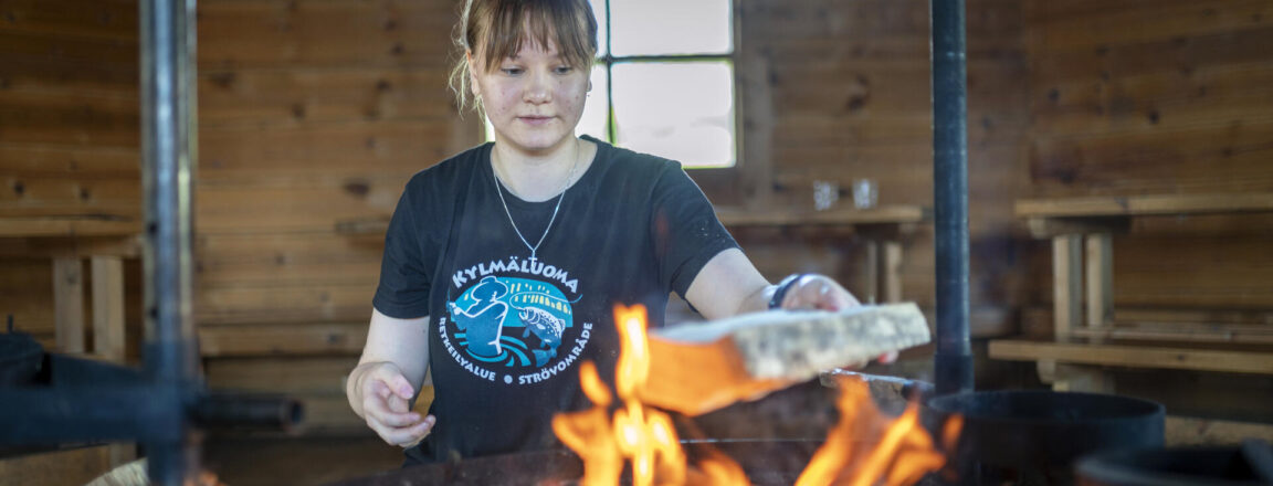 Nuori nainen lisää kodassa puita nuotioon. Hänellä on yllään musta t-paita, jossa on valkoinen ja sininen logo. Taustalla näkyy puinen pöytä ja penkki.