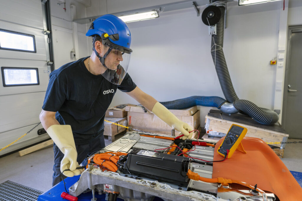 Henkilö työskentelee sisätiloissa kypärä ja suojalasit päässään, tutkien sähköajoneuvon akkua. Hän käyttää mittaria ja suojaavia hanskoja.