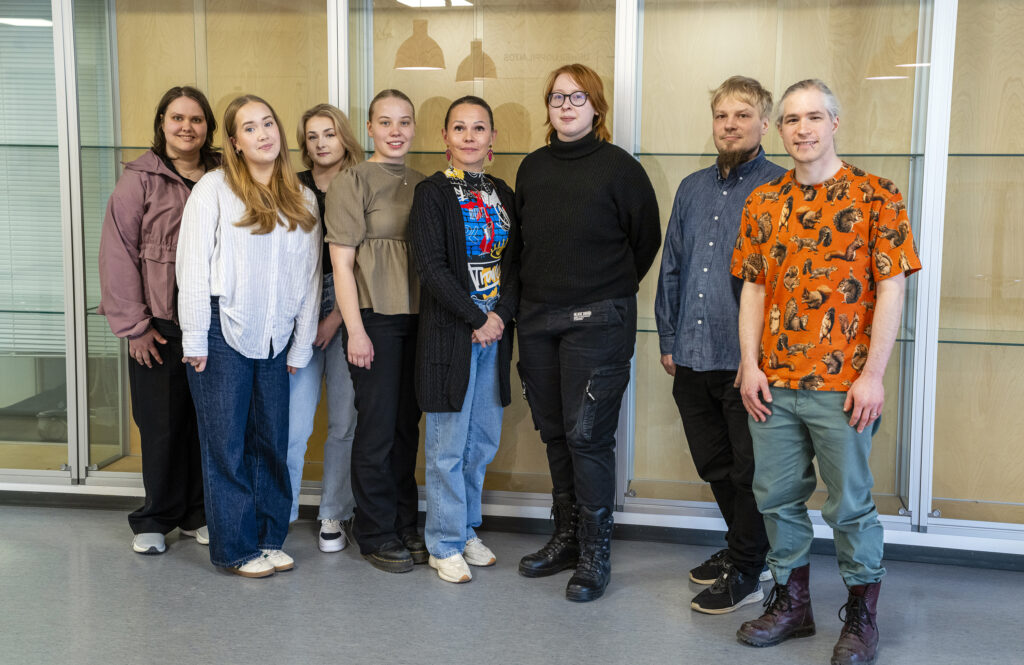 Kahdeksan henkilöä seisoo rivissä sisätiloissa, taustalla on lasivitriini. Henkilöt ovat erikokoisia ja -näköisiä, pukeutuneina erilaisiin vaatteisiin.