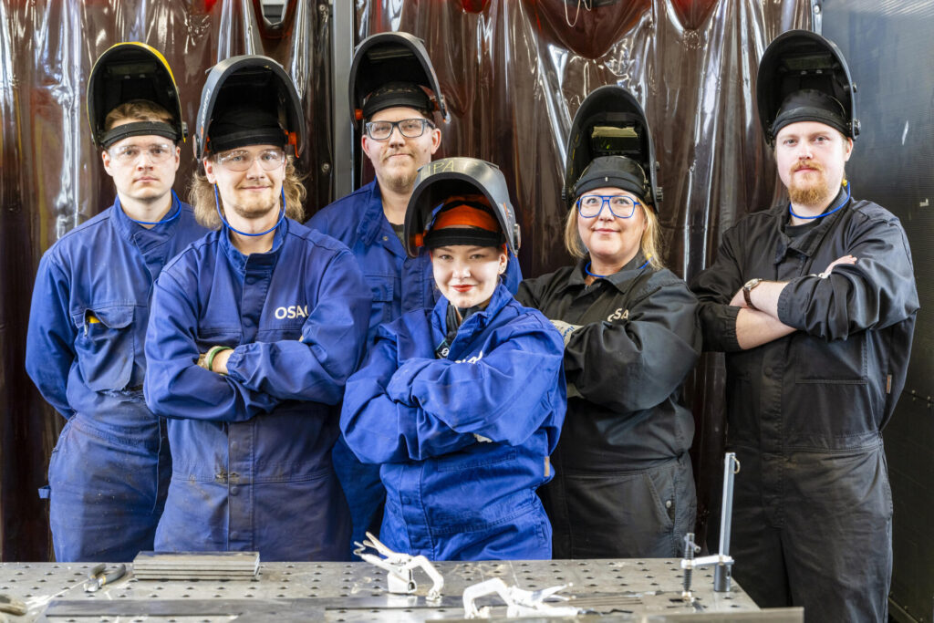 Ryhmä henkilöitä seisoo yhdessä työvaatteissaan ja hitsauskypärissään; etualalla on metallinen työpöytä, jonka päällä on työvälineitä.