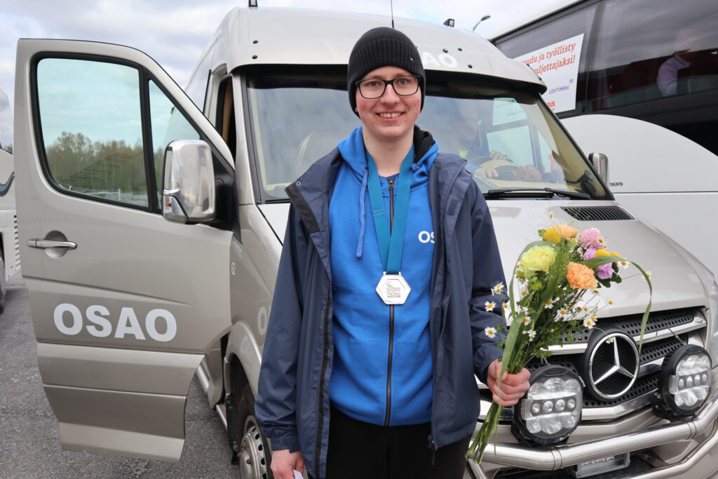 Nuori henkilö seisoo OSAO-logolla varustetun pakettiauton edessä. Hänellä on päällään sininen huppari ja musta pipo, ja kaulassaan hopeamitali. Hän pitää kädessään kukkakimppua ja hymyilee.