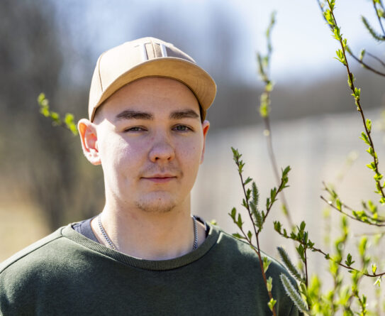 Nuori mies seisoo ulkona auringonpaisteessa. Hänellä on päällään vihreä paita ja beige lippalakki. Hänen takanaan on sumea tausta ja vihreitä oksia näkyy etualalla.