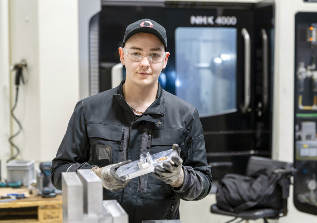 Henkilö seisoo teollisuusympäristössä ja pitää metallista komponenttia käsissään. Hänellä on yllään suojalasit, hanskat ja musta työasu. Taustalla näkyy suurikokoinen kone.