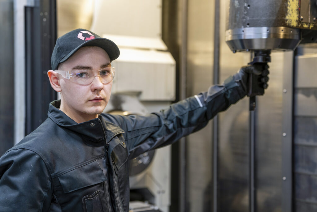 Henkilö seisoo teollisessa ympäristössä suojalaseihin ja lippalakkiin pukeutuneena. Hän pitää kädessään suurta metallista laitetta. Taustalla näkyy koneita ja metallirakenteita.