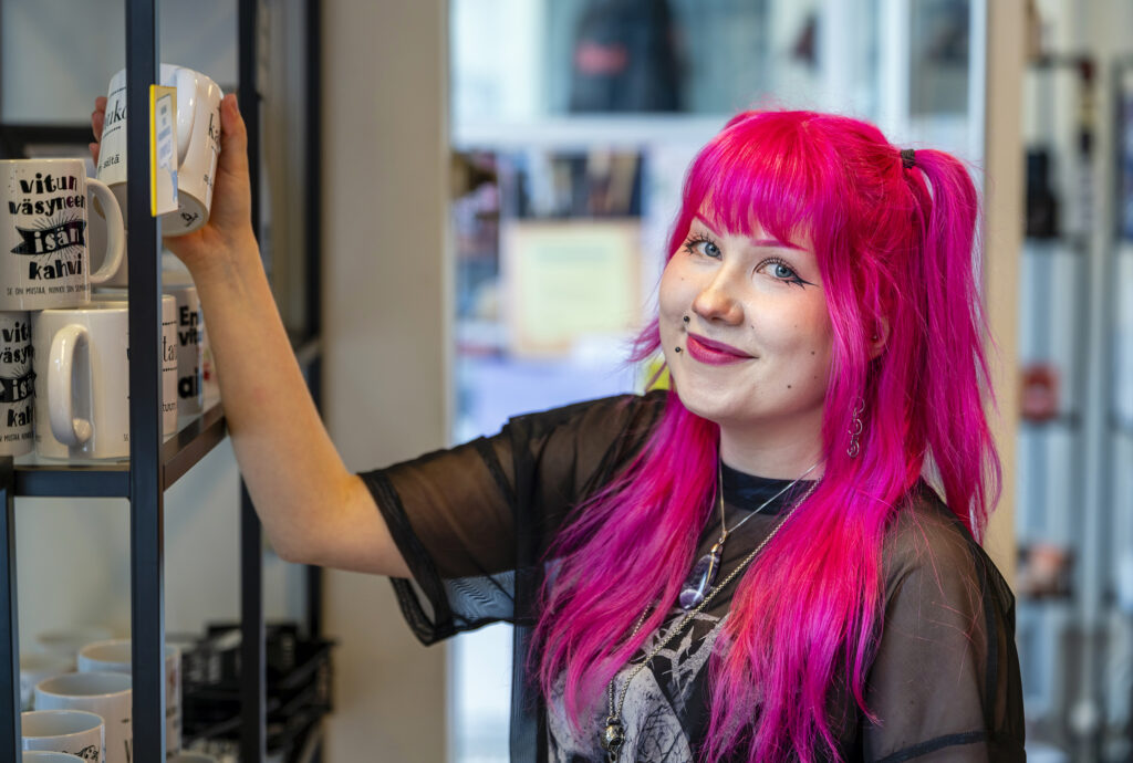 Henkilö, jolla on kirkkaan pinkit hiukset, seisoo hyllyn vieressä ja pitelee mukia. Hän hymyilee kameralle. Hyllyllä näkyy useita mukeja, joissa on tekstiä.