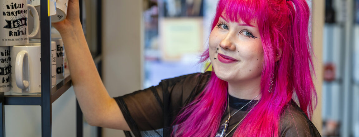Henkilö, jolla on kirkkaan pinkit hiukset, seisoo hyllyn vieressä ja pitelee mukia. Hän hymyilee kameralle. Hyllyllä näkyy useita mukeja, joissa on tekstiä.