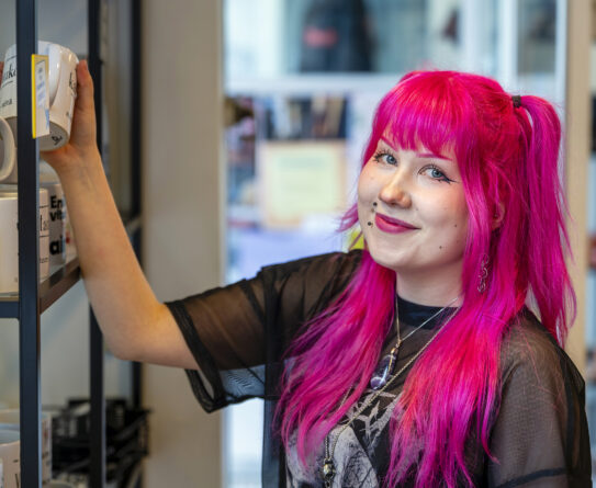 Henkilö, jolla on kirkkaan pinkit hiukset, seisoo hyllyn vieressä ja pitelee mukia. Hän hymyilee kameralle. Hyllyllä näkyy useita mukeja, joissa on tekstiä.