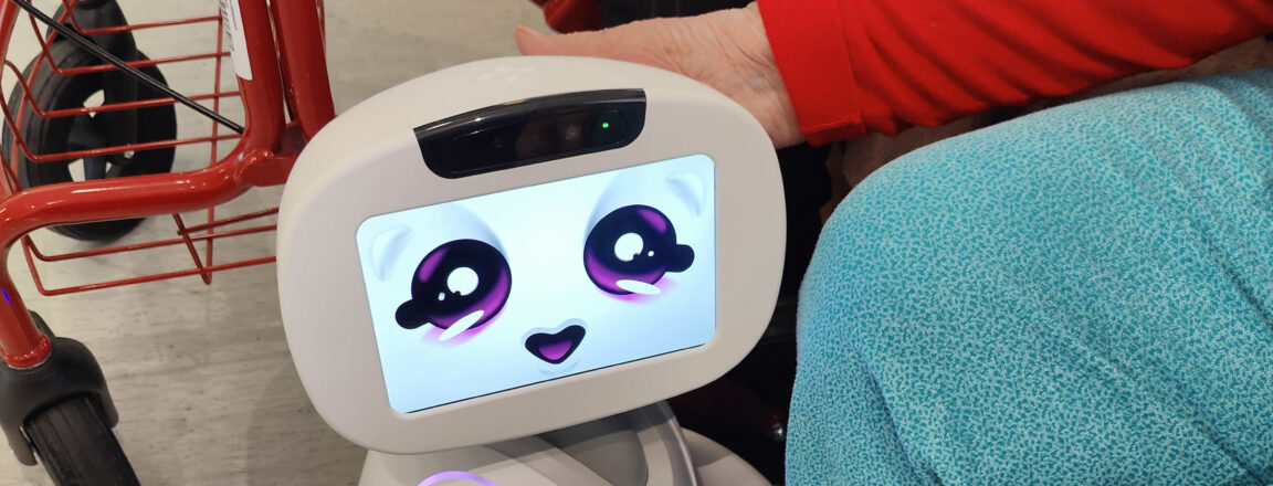 Henkilö istuu sohvalla ja koskettaa suloista robottia, jonka näytöllä on hymyilevät kasvot. Taustalla näkyy punainen rollaattori.