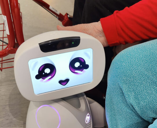Henkilö istuu sohvalla ja koskettaa suloista robottia, jonka näytöllä on hymyilevät kasvot. Taustalla näkyy punainen rollaattori.