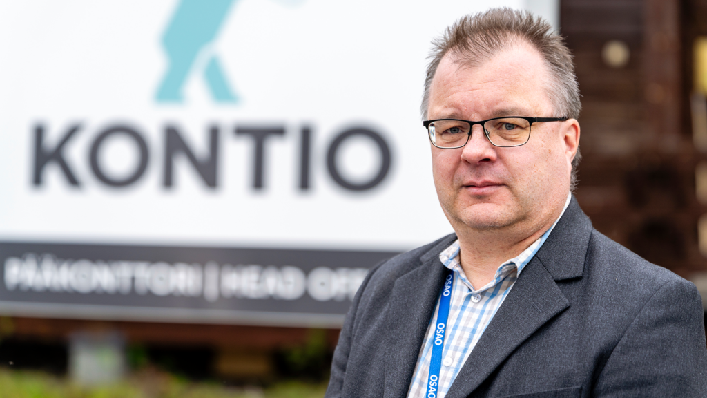 Mies seisoo ulkona kyltin edessä, jossa lukee 'Kontio Pääkonttori'. Hänellä on päällään tumma puku ja silmälasit, ja hän katsoo kameraan.