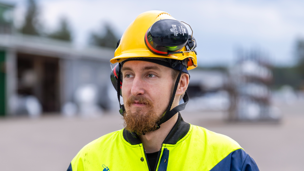 Mies, jolla on keltainen suojakypärä ja kirkkaan keltainen työtakki. Hänellä on myös kuulosuojaimet kypärän päällä. Taustalla näkyy epätarkka ulkoympäristö, teollisuusalue.