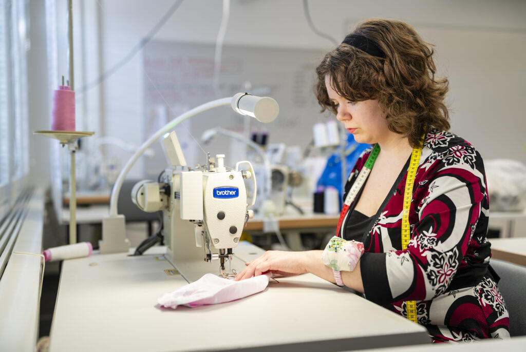 Nainen istuu ompelukoneen ääressä ja ompelee kangasta. Hänellä on värikäs paita ja mittanauha kaulassa. Taustalla näkyy muita ompelukoneita ja vaaleat seinät.